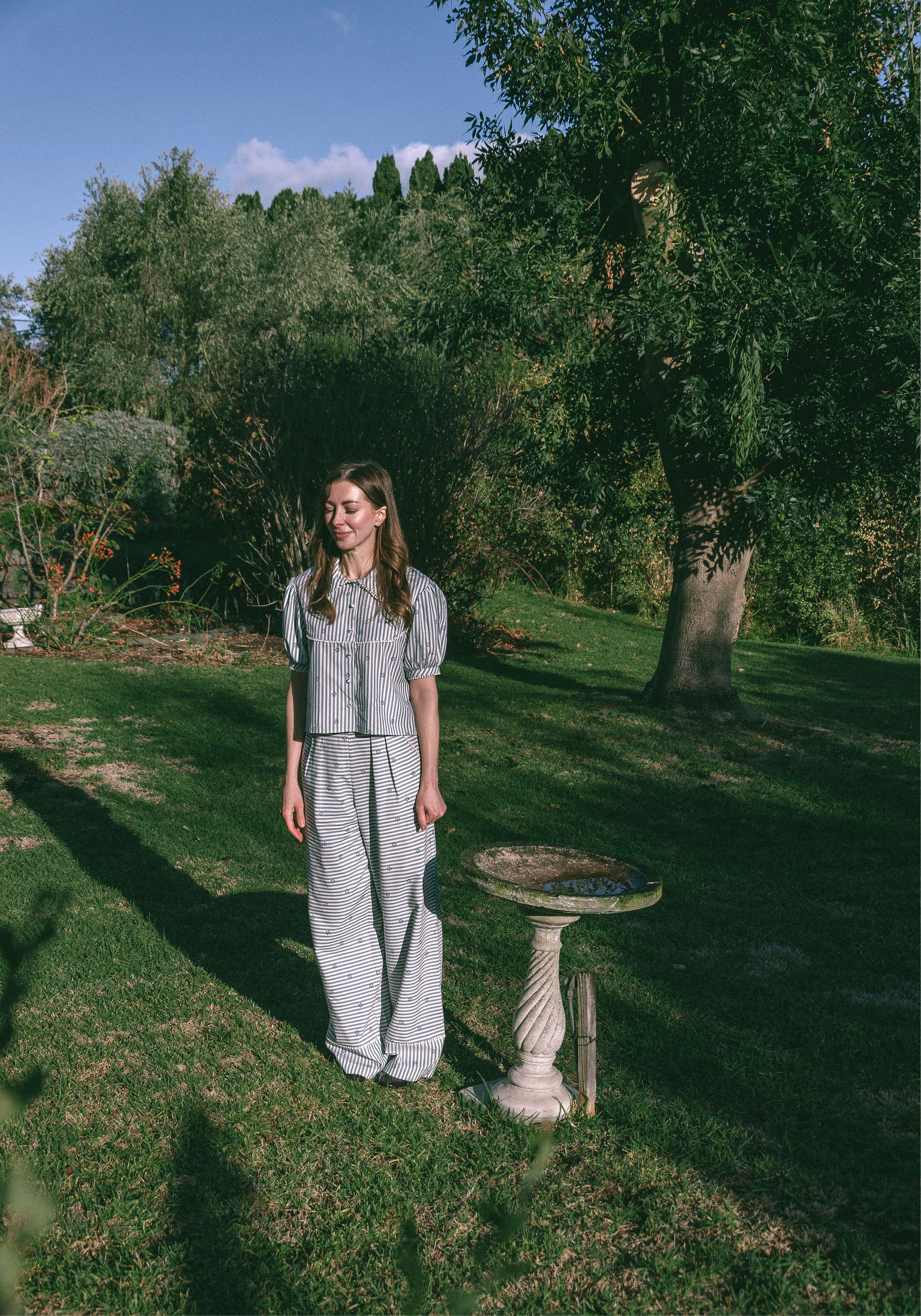 Young lady in a blue and white stripe pyjama set standing in a garden with trees and a bird bath.