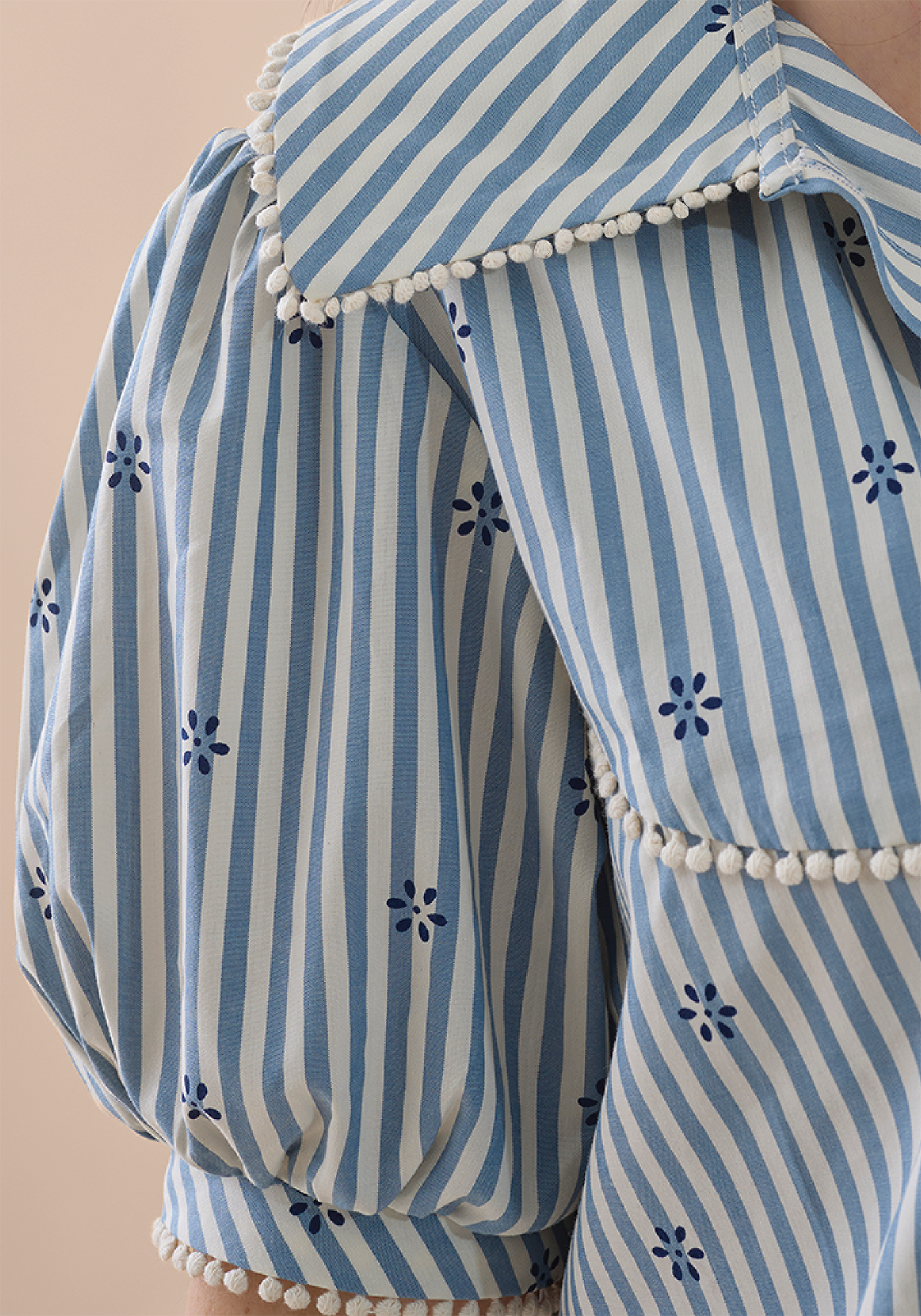 Close up of blue and white striped blouse garment with floral patterns and pom-pom details on a beige background